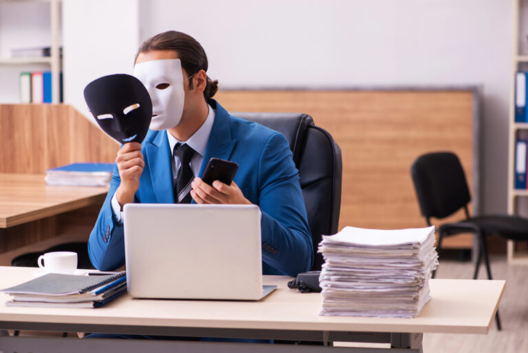 Businessman in a blue suit wears a white mask while holding another mask and looking at his phone at an office desk with a laptop.