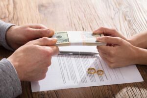 Two people exchange a bundled stack of cash over a signed document and rings on a wooden table, implying a financial agreement or settlement.